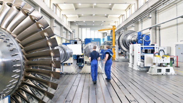Zwei Personen befinden sich in einer betrieblichen Werkstätte und sind umgeben von Maschinen.